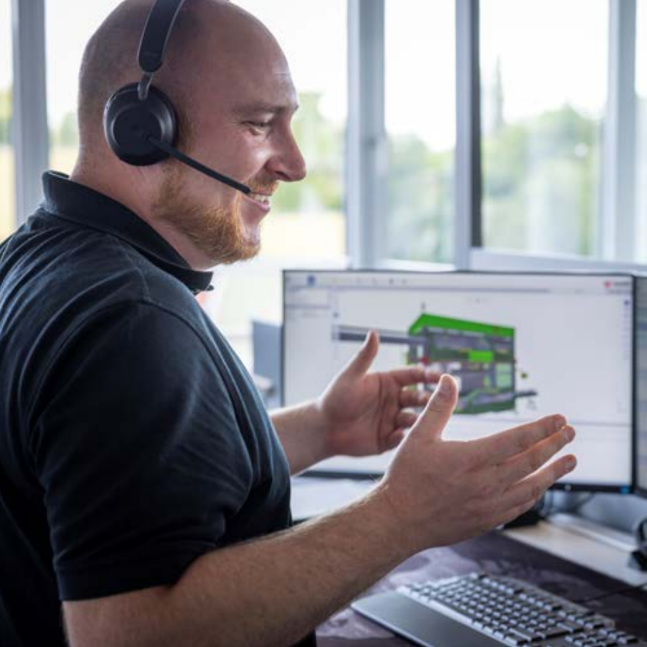 Technical expert providing remote customer support via headset while interacting with a digital machinery interface displayed across three monitors.