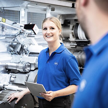 A woman is standing in front of machine and looks at a man who is also standing in front of machine.