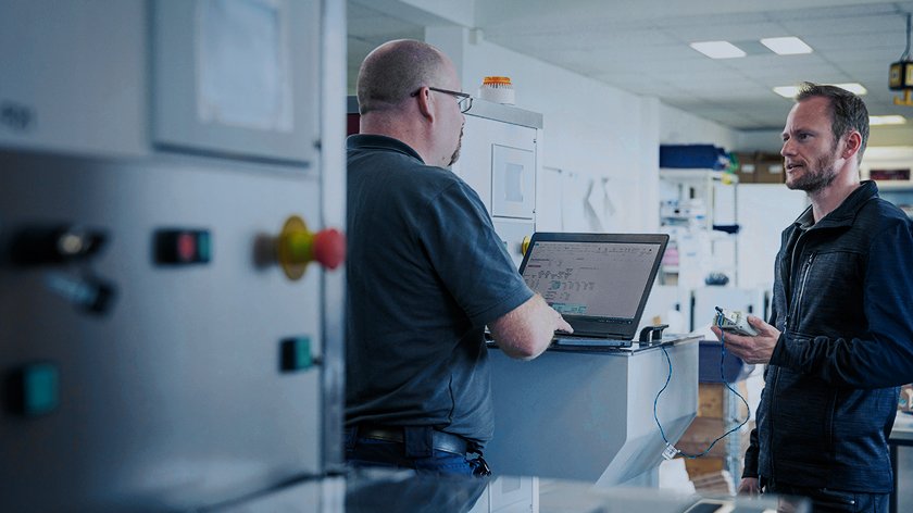 Two men standing next to a machine and talking while the man on the left is running a diagnostics program on a laptop.