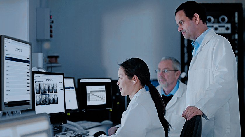 A woman and man are sitting and looking at multiple screens, showing graphs, while another man is standing behind the woman.