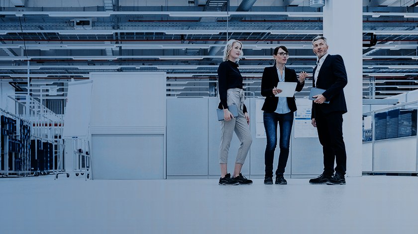 Two women and a man with files are standing in an empty area of a factory. The woman in the middle is explaining something. 