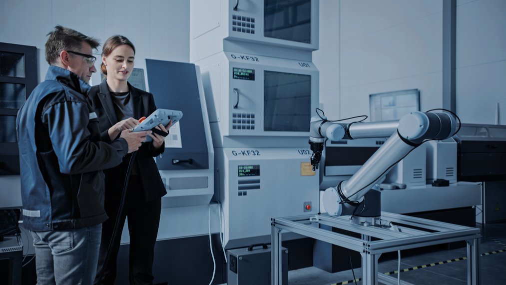 A man and a woman are holding a tablet and talking. They are standing in front of a machine and left of a robotic arm. 