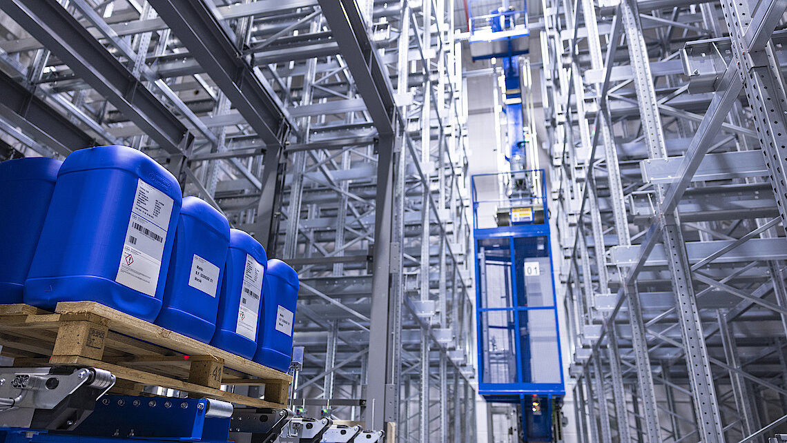 Photograph of Körber’s new automated high-rack warehouse with blue barrels on euro-pallets in the left corner. 