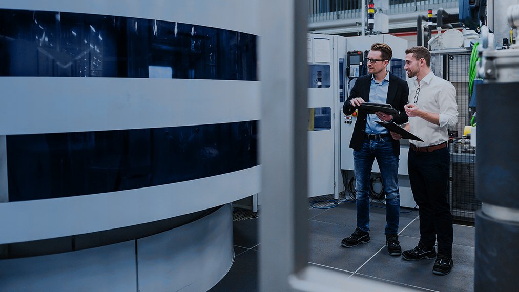 Two men are discussing and standing in front of a machine, one is holding a tablet, the other a clipboard. 