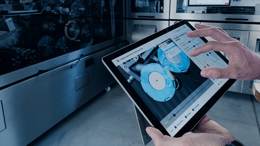 Zwei Hände halten und benutzen ein Tablet, welches Diagnosen und Steuerungen zeigt. Im Hintergrund steht eine Maschine.