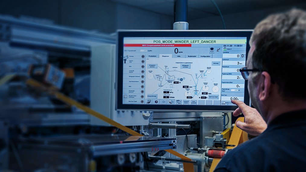 Ein Mann betrachtet Produktionszahlen auf einem Kontrollbildschirm, der mit einer linksstehenden Maschine verbunden ist.