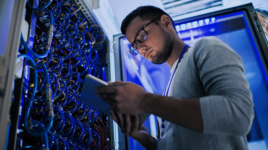 A server door is open and a man is standing in front of the server whilst looking down at a tablet he is holding.