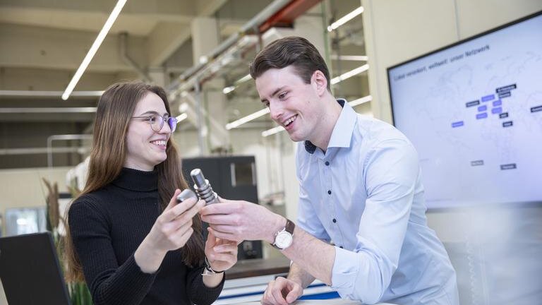 Zwei duale Studenten von Körber Technologies betrachten gemeinsam ein Werkstück