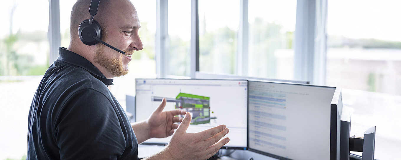 Technical expert providing remote customer support via headset while interacting with a digital machinery interface displayed across three monitors.